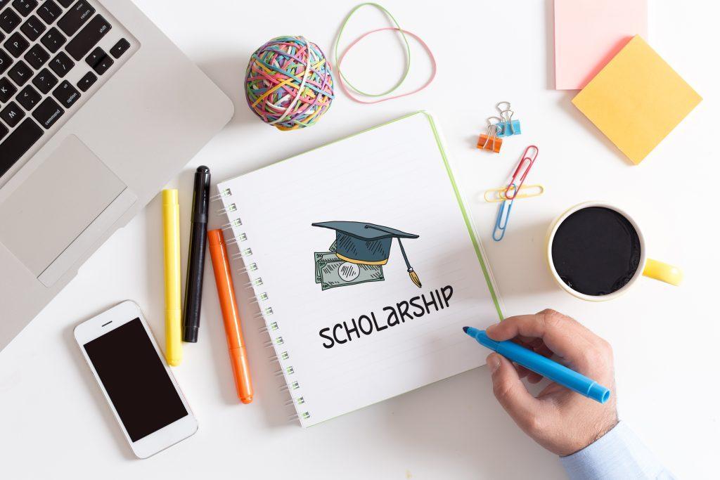 notebook on a messy desk with a drawing of a cap with cash and the word scholarship